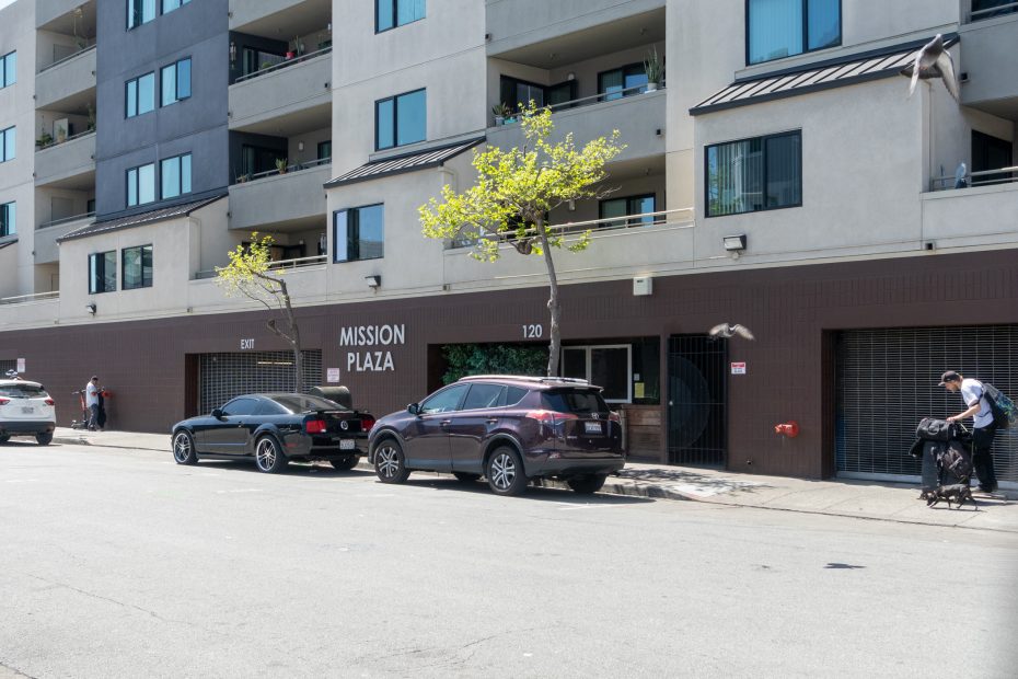 Street view of Mission Plaza building with parked cars and a person pushing a stroller. Birds are in mid-flight.