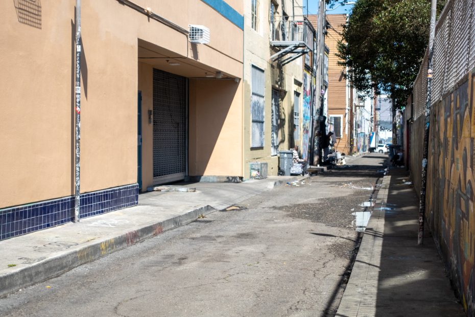 Narrow urban alley with beige and tan buildings, graffiti on walls, and scattered debris. A tree shades part of the street.