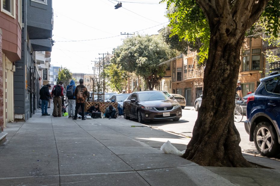 A group of people gather on a city sidewalk beside a parked car, near a tree. Urban buildings and utility lines are in the background.