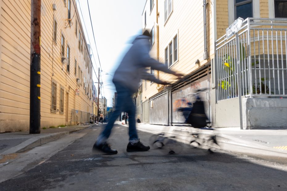 A blurred person pushes a shopping cart quickly through a narrow urban alley with yellow buildings on a sunny day.