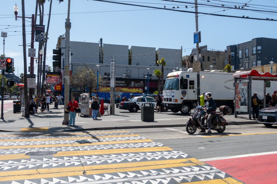 Urban intersection with pedestrians, a police vehicle, and a motorcycle. Buildings and a street market are in the background. Crosswalk features geometric patterns.