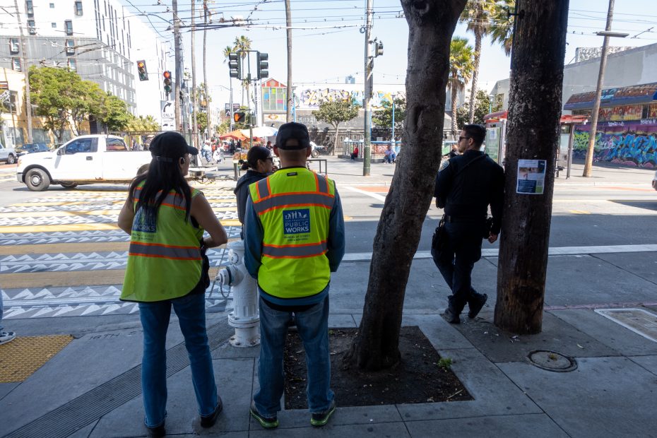 People wearing high-visibility vests stand near a street corner with trees and traffic in the background. A person in black clothing walks nearby.