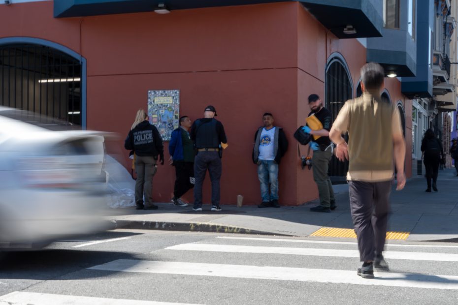 Police officers speak with three individuals on a sidewalk near a building as a car passes and a person walks nearby.