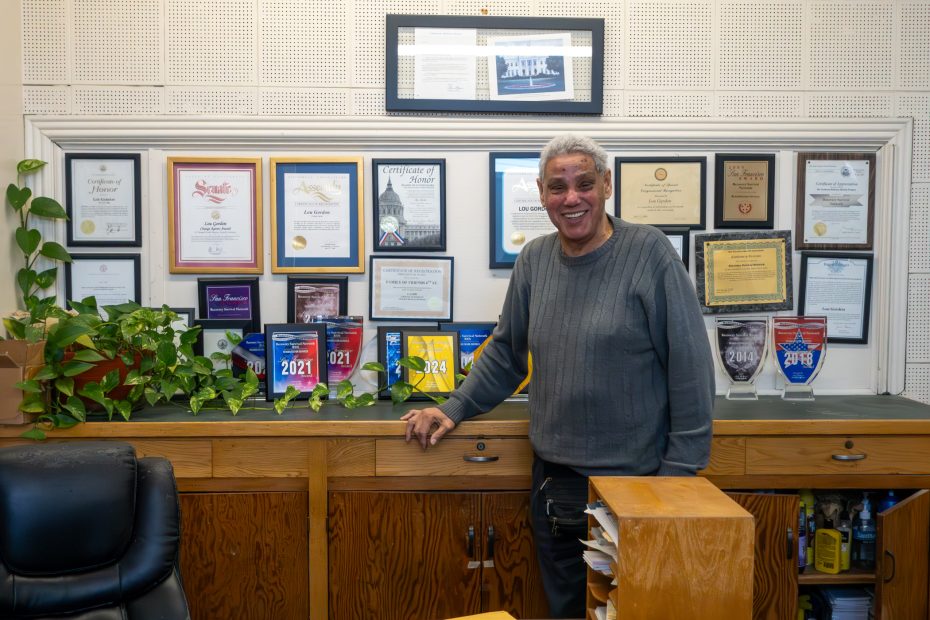A person stands smiling in front of a wall filled with awards and certificates, next to a countertop displaying multiple award plaques and plants.