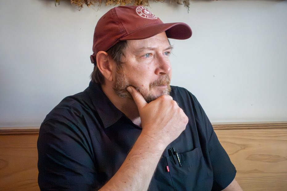 Man in a red cap and black shirt rests his chin on his hand, sitting indoors. Brown hair and facial hair, with pens in his shirt pocket, looking to the side thoughtfully.