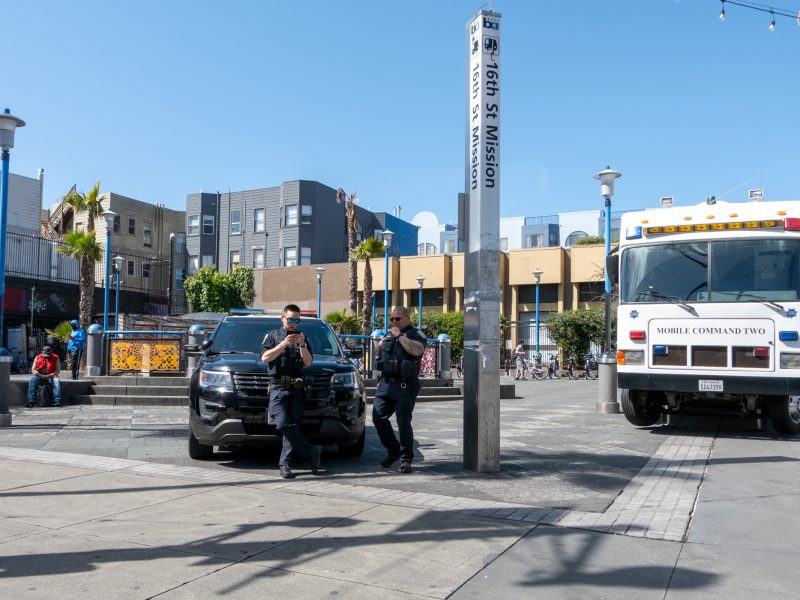 Two police officers stand beside a patrol vehicle at the 16th St. Mission station. A Mobile Command vehicle is parked nearby. Urban setting with trees and buildings in the background.