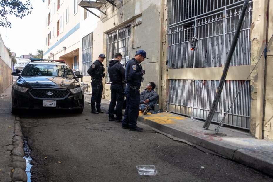 Police officers standing near a patrol car interact with a seated individual beside a gated entrance in an alley.