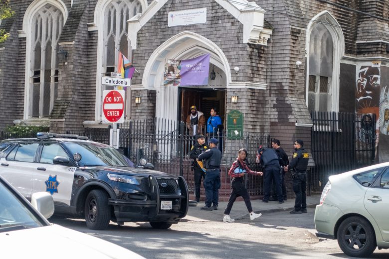 Police officers and people outside a gray stone building with arched windows. A police car is parked nearby, and a "Do Not Enter" sign is visible.
