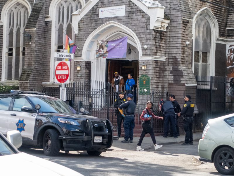 Police officers and people outside a gray stone building with arched windows. A police car is parked nearby, and a "Do Not Enter" sign is visible.