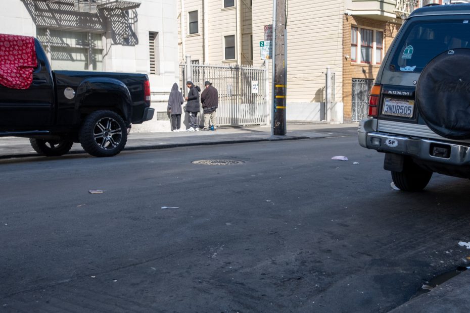 Two parked trucks on a street; three people stand on the sidewalk in conversation.