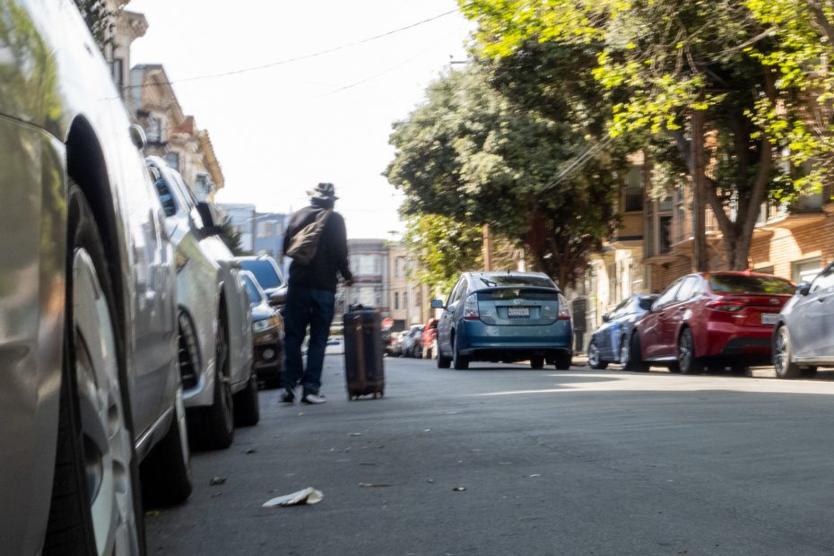A person rolls a suitcase up a sunlit urban street lined with parked cars and trees.