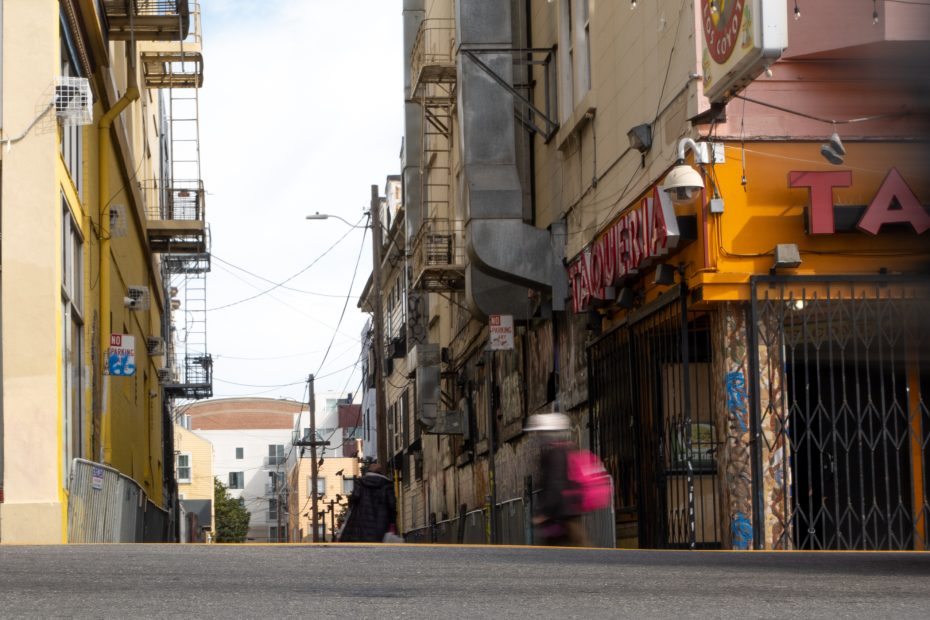 Urban street scene with blurred pedestrians walking past buildings adorned with signs and fire escapes. Overhead wires stretch across a cloudy sky.
