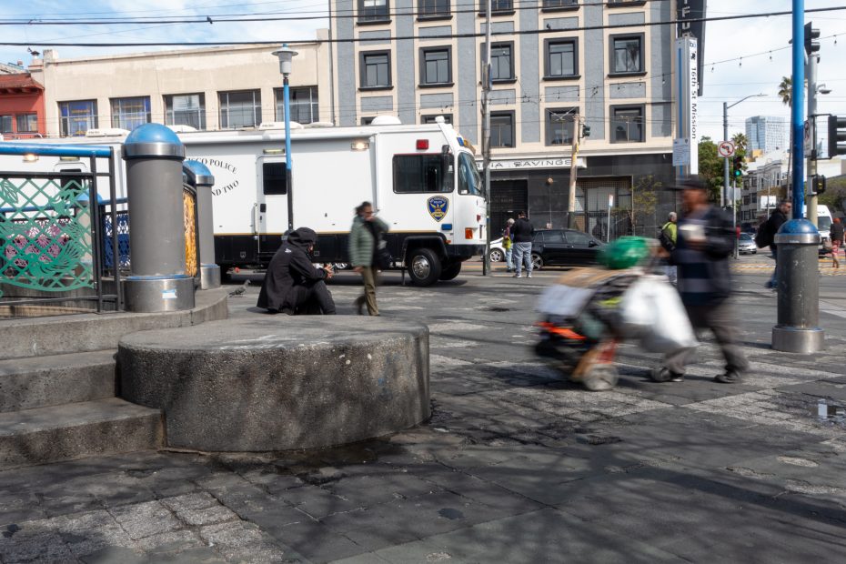 A city street scene showing a person sitting on steps, a blur of someone pushing a cart with bags, and a police truck parked on the road in front of a large building.
