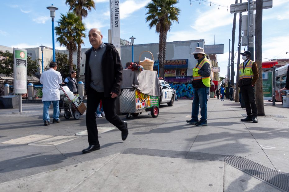 A busy street scene with pedestrians walking, two workers in reflective vests, palm trees, and a person pushing a cart with items. A mural is visible on the building in the background.