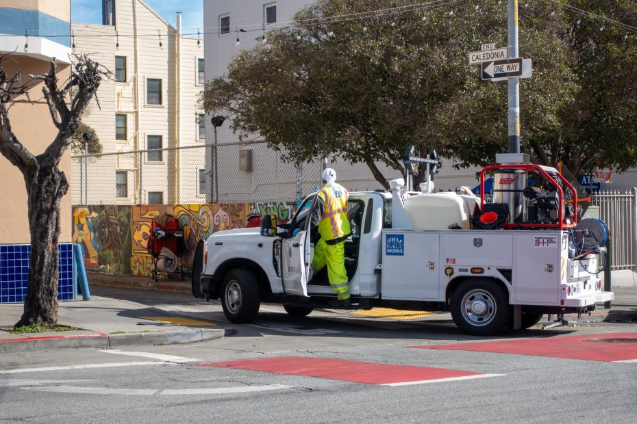 Utility worker in a bright vest and helmet stands on a truck parked at a street corner, adjacent to a vibrant mural and leafless trees.