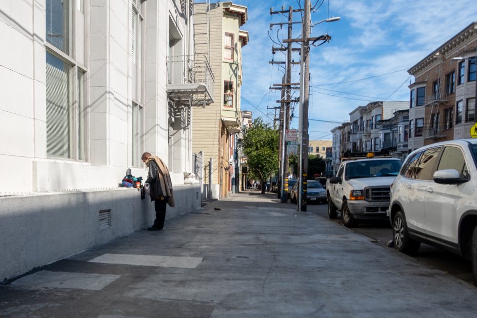 Person wearing a coat standing on a sidewalk next to a white building, looking at objects on a ledge. Street lined with parked cars and buildings. Clear blue sky above.