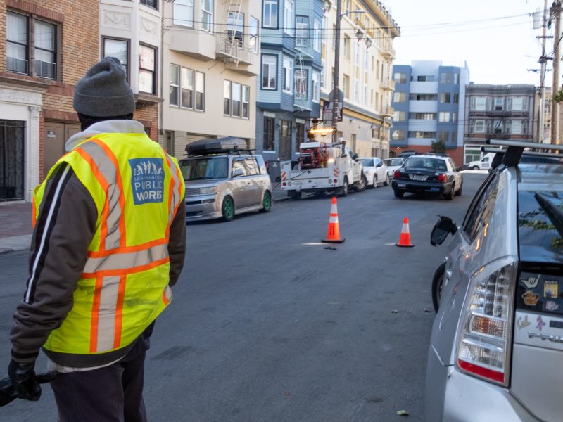 A person in a "Public Works" vest stands on a city street near a row of parked cars and a tow truck, with traffic cones placed on the road.