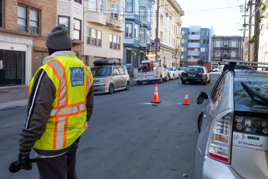 A person in a "Public Works" vest stands on a city street near a row of parked cars and a tow truck, with traffic cones placed on the road.