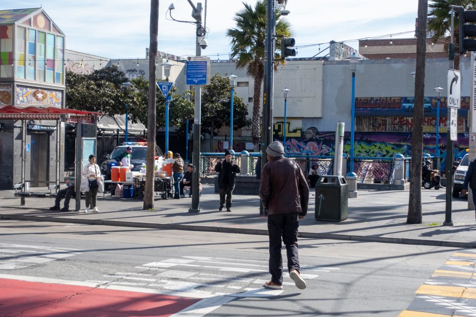A person in a jacket and beanie crosses a street in an urban area, with others near a food stand and colorful graffiti in the background.