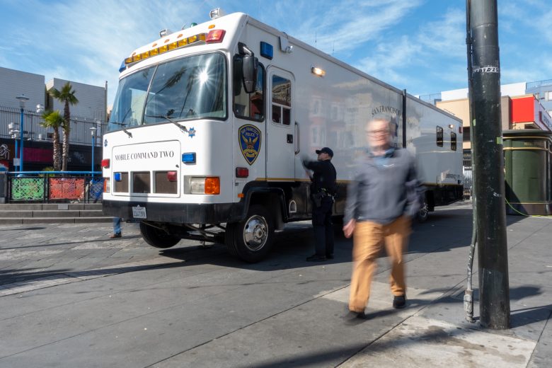 A large, white mobile command vehicle is parked on a street. Two officers stand by the vehicle, while a person walks past in the foreground, appearing blurred. Buildings are in the background.