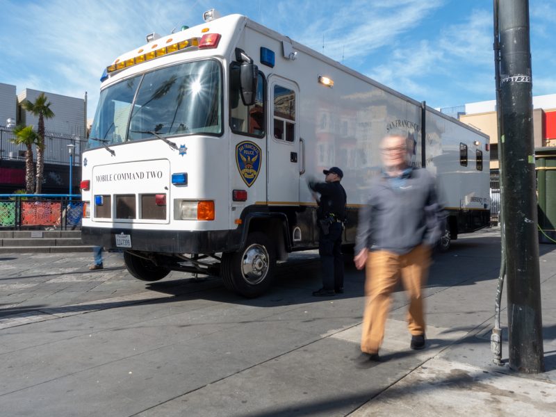 A large, white mobile command vehicle is parked on a street. Two officers stand by the vehicle, while a person walks past in the foreground, appearing blurred. Buildings are in the background.