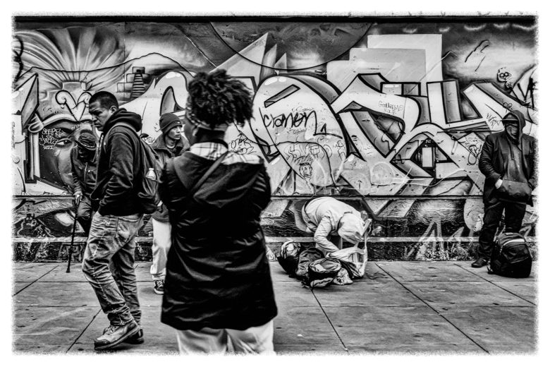A group of people stand and walk on a city sidewalk in front of a wall covered with graffiti; one person appears to kneel over a bag. The image is in black and white.
