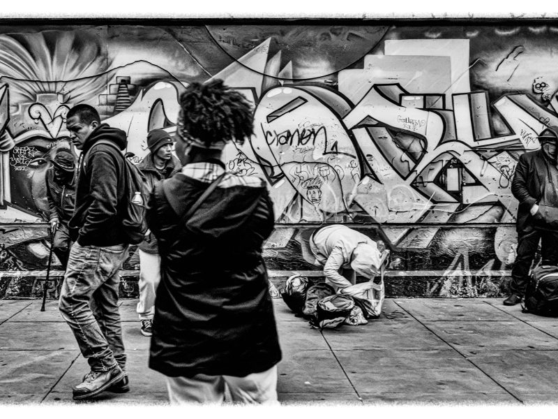 A group of people stand and walk on a city sidewalk in front of a wall covered with graffiti; one person appears to kneel over a bag. The image is in black and white.