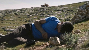 A person in a blue shirt lies on the ground outdoors with head resting on a rock; construction vehicle visible in the background.