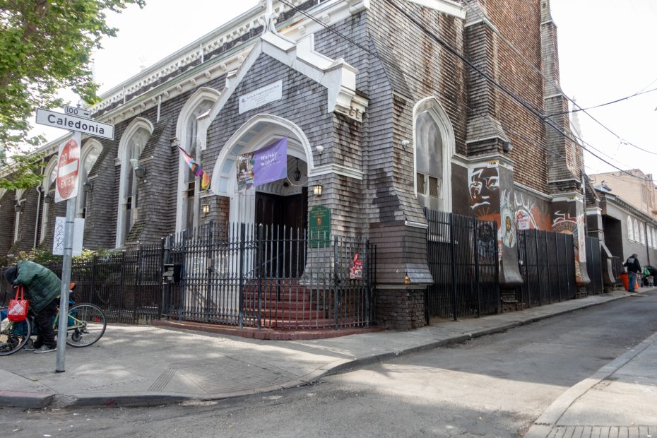 A large brick church building with arched windows, a gated entrance, flags, and murals on the walls, located at the corner of Caledonia Street.
