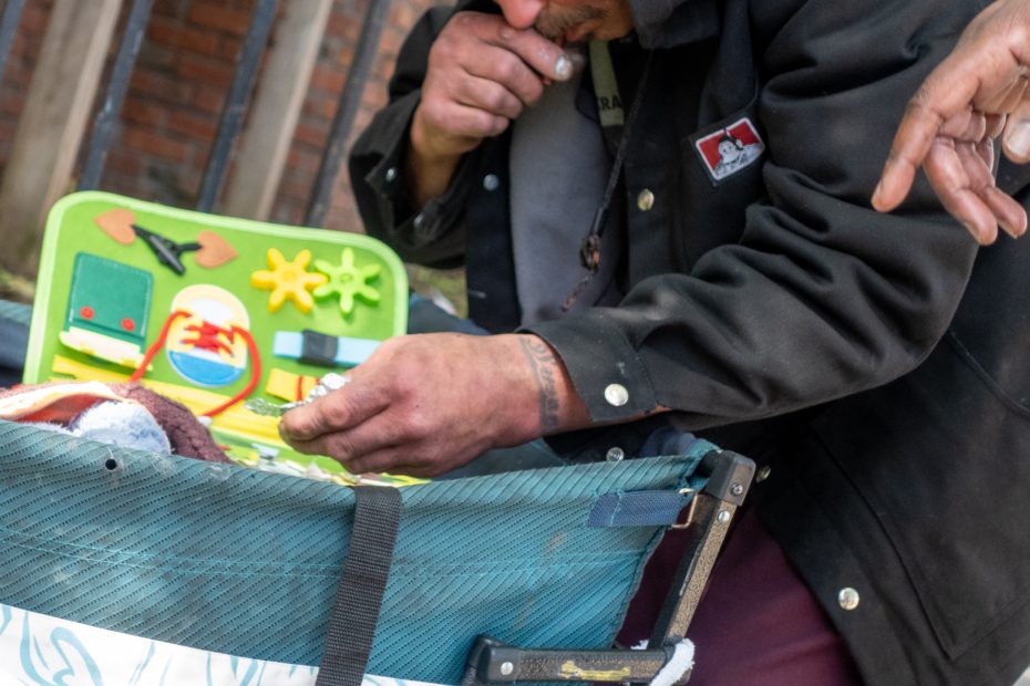 A person with dirty hands leans over a blue carrier, looking at a green tray with colorful plastic toys and items, while another hand reaches in from the side.