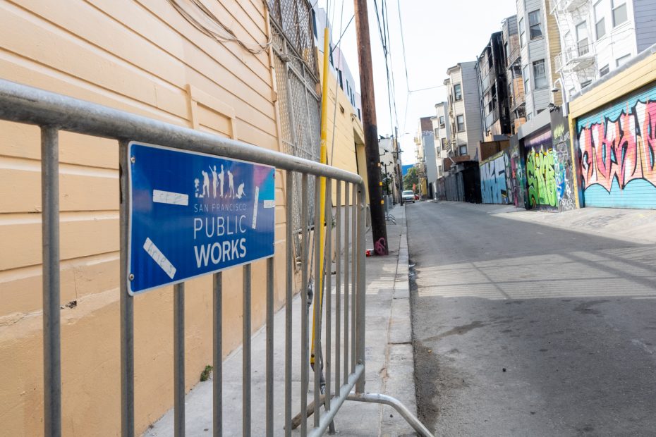 A metal barricade with a San Francisco Public Works sign blocks access to an alley lined with buildings and graffiti.