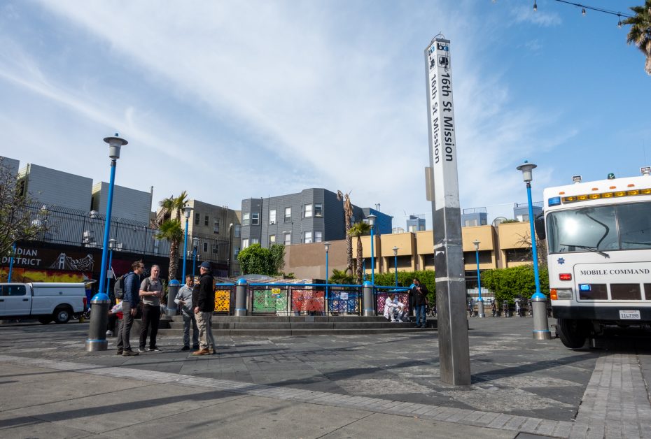 Outdoor plaza at 16th St Mission BART station, with people talking, colorful murals, and a mobile command vehicle parked nearby under a clear sky.