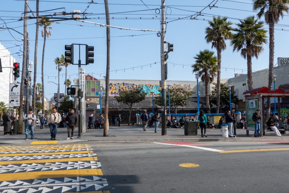 People wait at a crosswalk on a city street lined with palm trees, street art, and overhead cables under a clear sky.