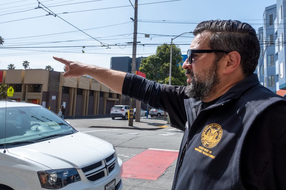A man wearing sunglasses and a black jacket with an official seal points across a city street near a white vehicle and a crosswalk.