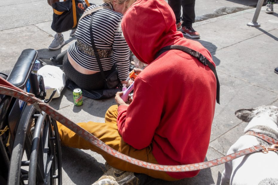 A person in a red hoodie and another person sit on a sidewalk near a wheelchair, a soda can, and a dog on a leash.