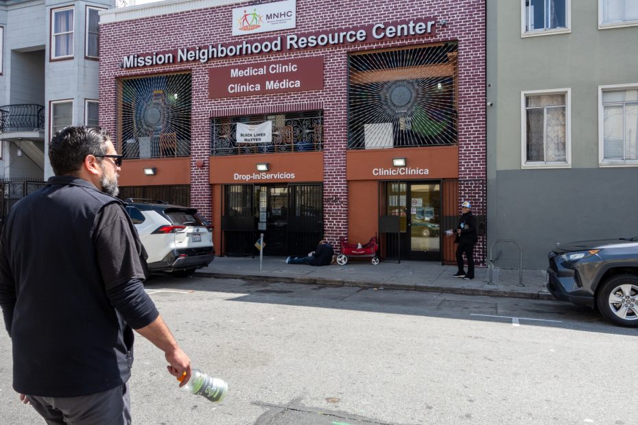 A man walks across the street toward the Mission Neighborhood Resource Center, which offers medical clinic and drop-in services; two people sit near the entrance.
