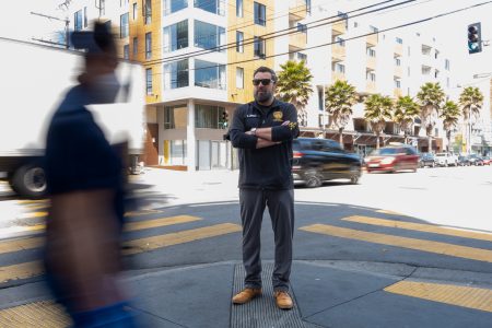 A man stands with arms crossed at a city intersection as cars and a blurred pedestrian move around him. Urban buildings and palm trees are in the background.