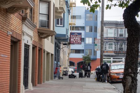 A sidewalk view of a city street with several people near belongings, a sign for Kaltash Hotel, and parked cars along the curb.