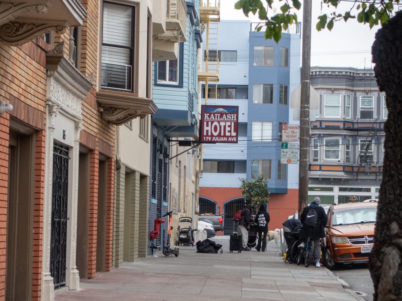A sidewalk view of a city street with several people near belongings, a sign for Kaltash Hotel, and parked cars along the curb.
