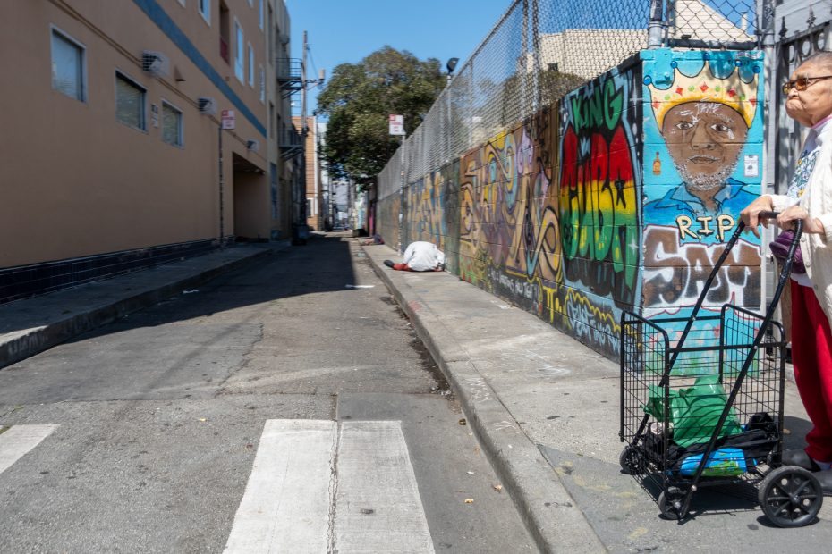 A street scene with graffiti on a wall. A person with a shopping cart stands on the right, and someone sits on the sidewalk in the distance. Buildings line the alley.