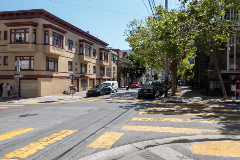 Street corner in an urban area with crosswalks, parked cars, and two-story buildings. Trees line the street, and a few people walk on the sidewalks.