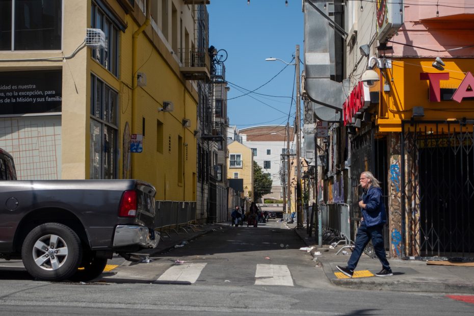 A person crosses an alley between a truck and a building with yellow walls. Shops are on the right, and a few people are visible in the background.