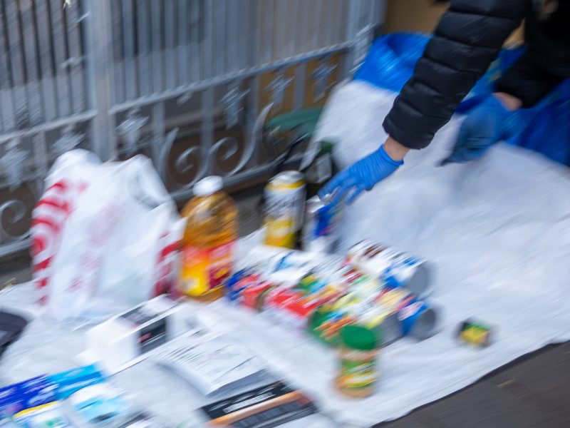 A person wearing gloves arranges various household items, including bottles and packages, on a tarp near a metal fence.