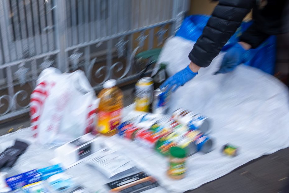 A person wearing gloves arranges various household items, including bottles and packages, on a tarp near a metal fence.