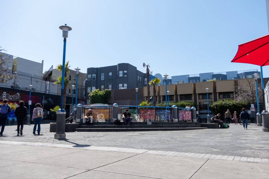 Urban plaza with people sitting and walking. Buildings in the background, red umbrella on the right. Clear blue sky.