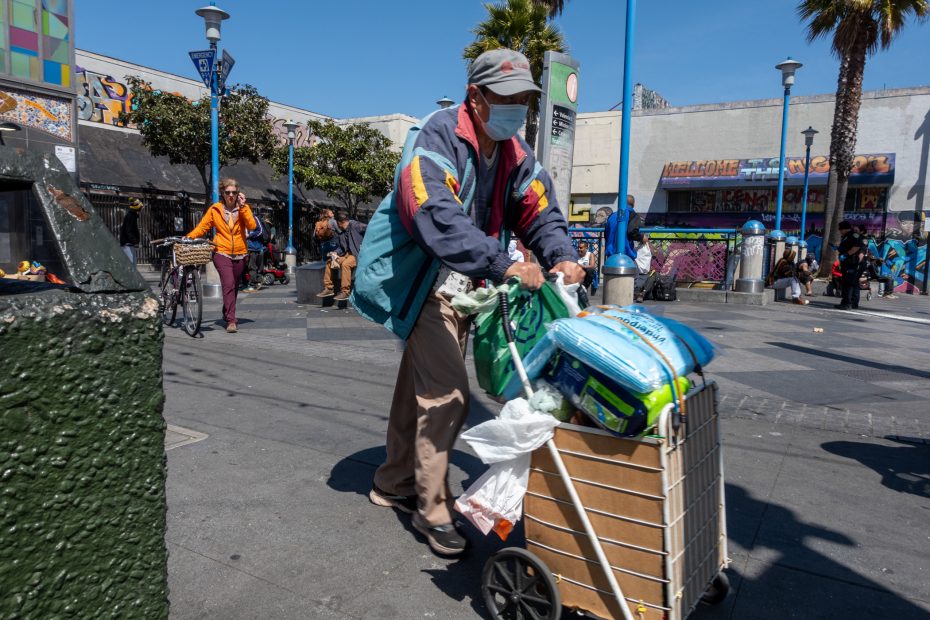 Person wearing a mask pushes a cart filled with bags on a busy urban street with graffiti and palm trees in the background.