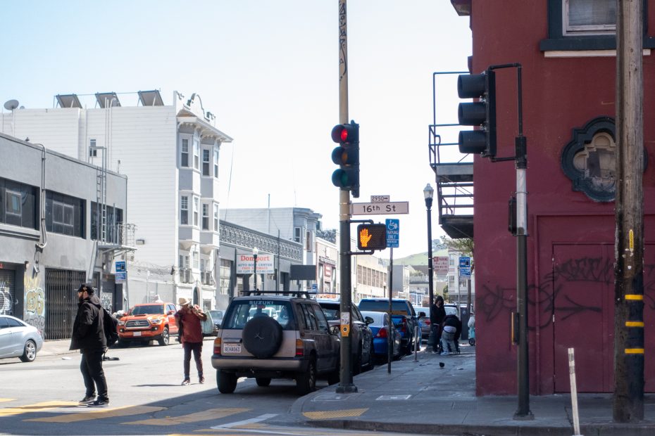 Urban street corner at 16th St with traffic lights and pedestrians. Vehicles are parked along the street, and a red building with graffiti is visible.
