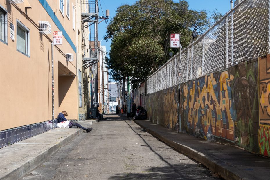 Urban alley scene with vibrant graffiti covering the right wall. A few people are sitting or lying on the ground along the left side. Sunny weather with shadows cast on the pavement.