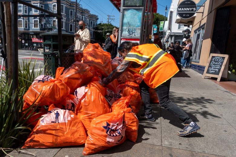 A person wearing an orange safety vest arranges a pile of orange garbage bags on a sidewalk near a storefront. Other pedestrians walk by on the urban street.