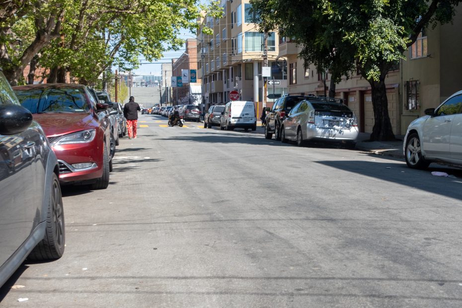 A street lined with parked cars on both sides, with a woman walking in the distance. Trees and multi-story buildings are visible alongside the road on a sunny day.